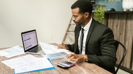 Finance professional in a suit reviewing financial documents with a laptop and calculator at a desk